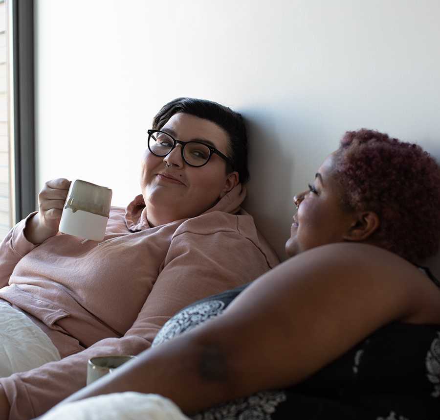 Two people sit in bed drinking coffee. They both have short hair and nose piercings.